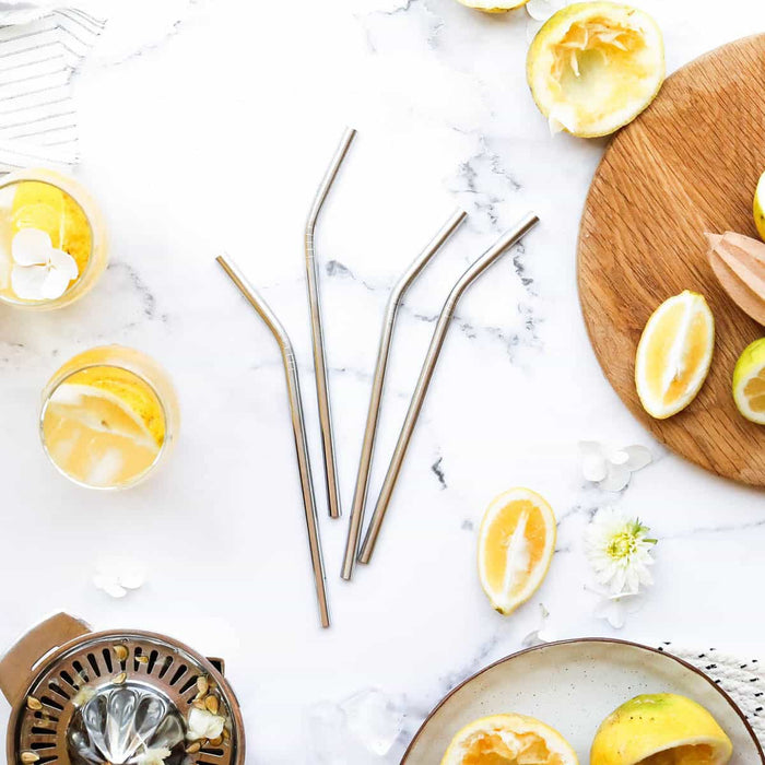 4 stainless steel straws elegantly laid out on a marble surface surrounded by a wooden cutting board, with sliced & juiced lemons & a glass of lemony drink.