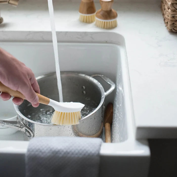 A hand uses a wooden-handled dish brush with a white bristle head to scrub inside a stainless steel pot in a white kitchen sink, with running water and soap visible.