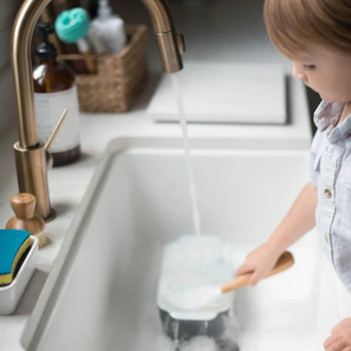 A young child in a light blue shirt stands at a white kitchen sink, washing a dark pot under running water from a brushed brass faucet. A blue sponge and soap dispenser are visible on the counter.