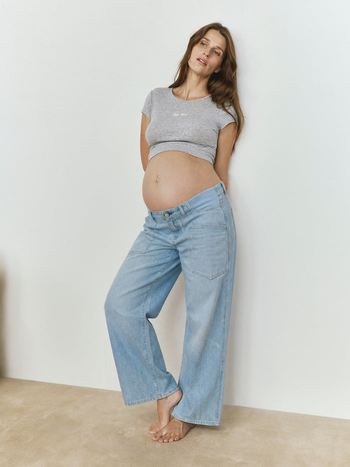 Pregnant woman wearing a gray crop top and blue jeans against a white wall.