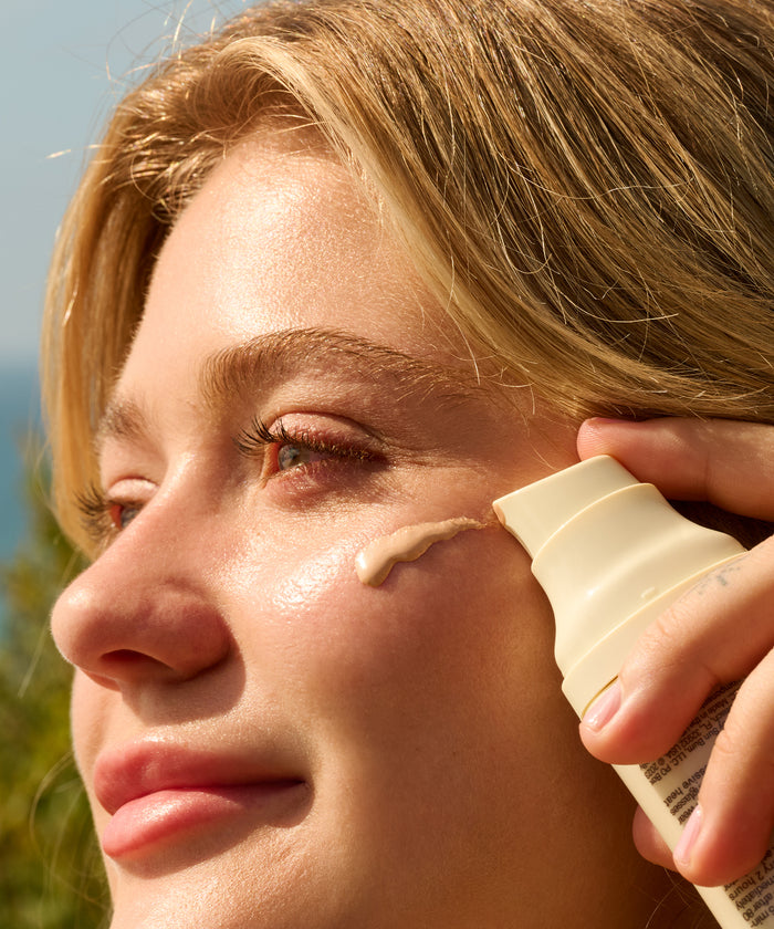 Close-up of a woman applying Daily Body + Glow Bundle cream by Sun Bum on her glowing cheek outdoors.
