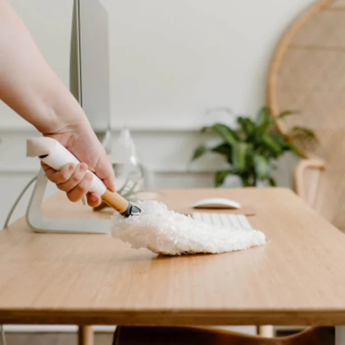 A hand holding a white microfiber duster with a wooden handle cleans a light wood desk in a bright, modern home office setting.