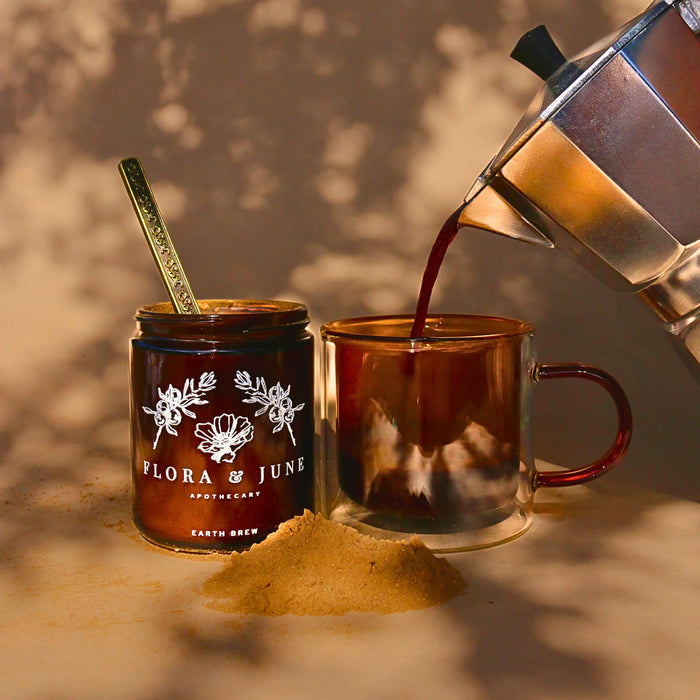 A soothing pour combining fresh coffee with Flora & June's Earth Brew into a clear mug beside its amber jar, with scattered herbal powder on a surface bathed in sunlight, capturing the essence of a healthful coffee substitute.