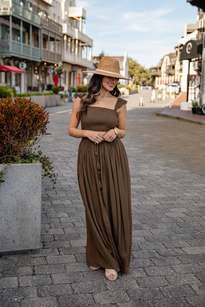 Woman in a brown dress and hat standing on a street with buildings and plants in the background