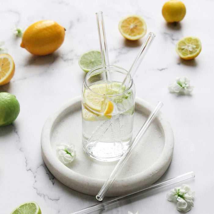 Ever Eco glass drinking straws styled in a glass of sparkling water with lemon and lime slices and flowers placed around. Sitting on a white coaster on a marble surface.
