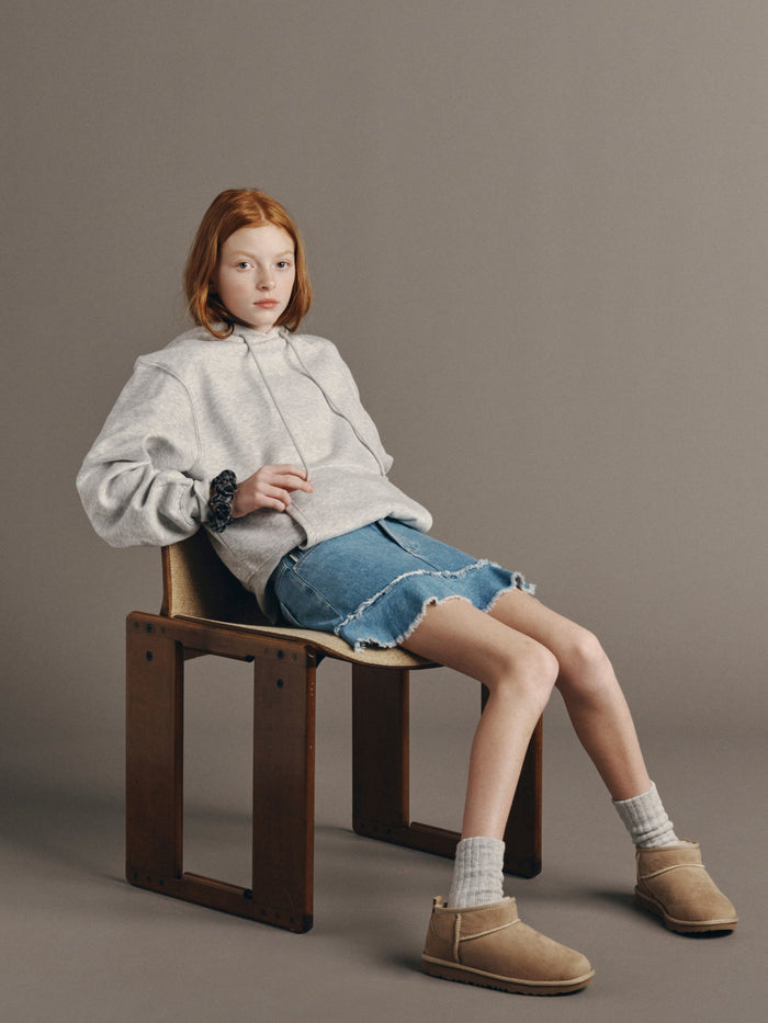 DL1961 girls' Jenny ruffle-hem mini skirt in light blue 'Unraveled' with a grey hoodie, seated on a wooden chair.