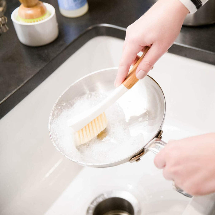 A close-up of a person's hands using a bamboo-handled dish brush with soft yellow bristles to clean a stainless steel pot inside a white sink filled with soapy water.