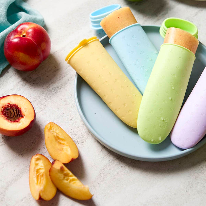 Colourful popsicle moulds filled with frozen fruit puree on a plate with peaches and peach slices on a light surface.