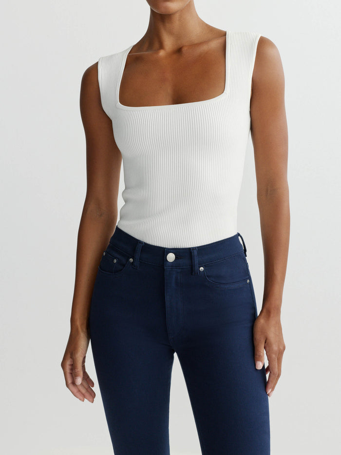 Person wearing a white sleeveless top and dark blue jeans on a light gray background