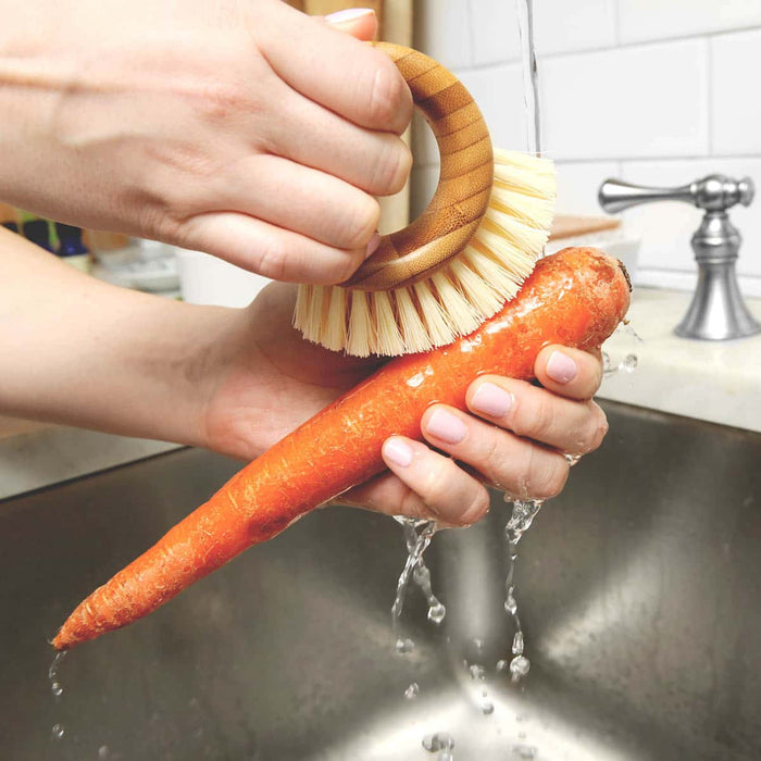 Person washing a carrot under running water with the ring veggie brush in a kitchen sink.